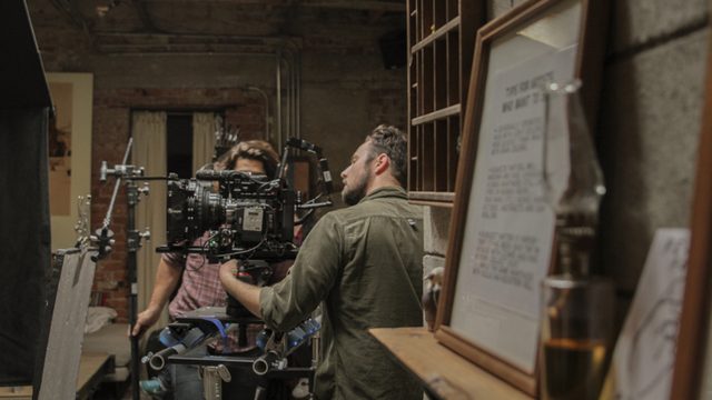 On set: two crew at a cinema camera on a rig in a brick basement.