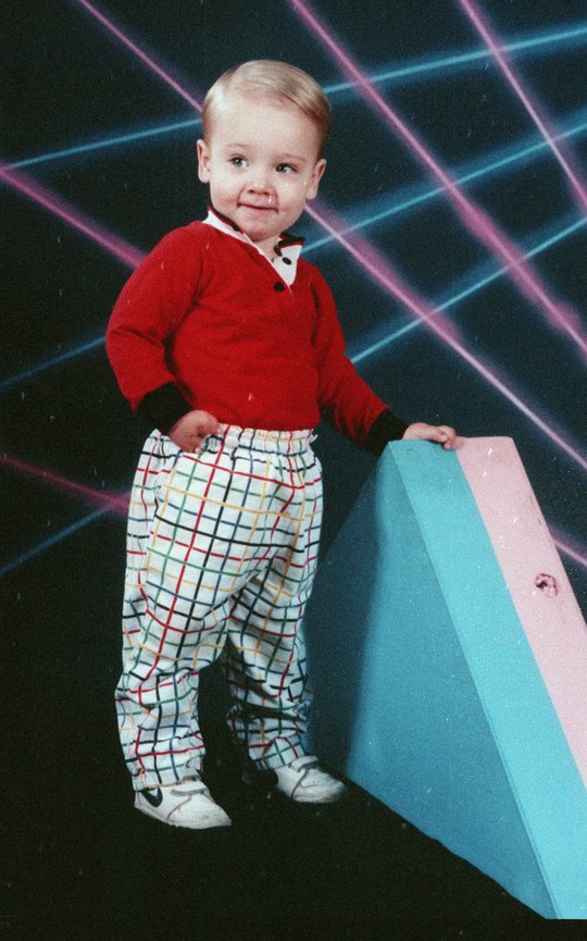 A toddler in a red shirt and plaid pants posing against a laser backdrop in a portrait studio.