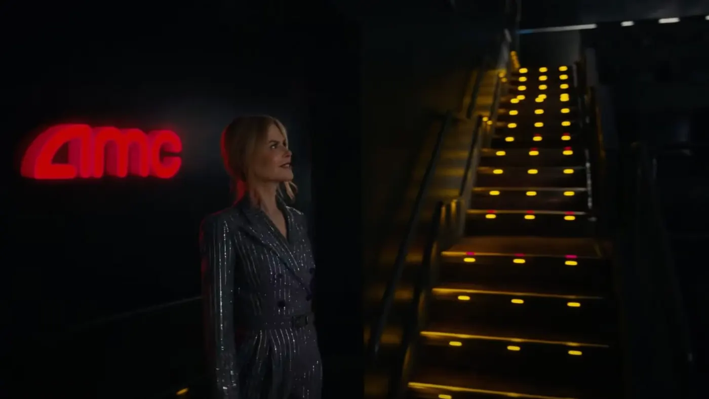 A frame from the AMC spot, dimmed: Nicole Kidman stands in a dark AMC lobby beside a red AMC sign, looking up a staircase lit with small yellow lights.